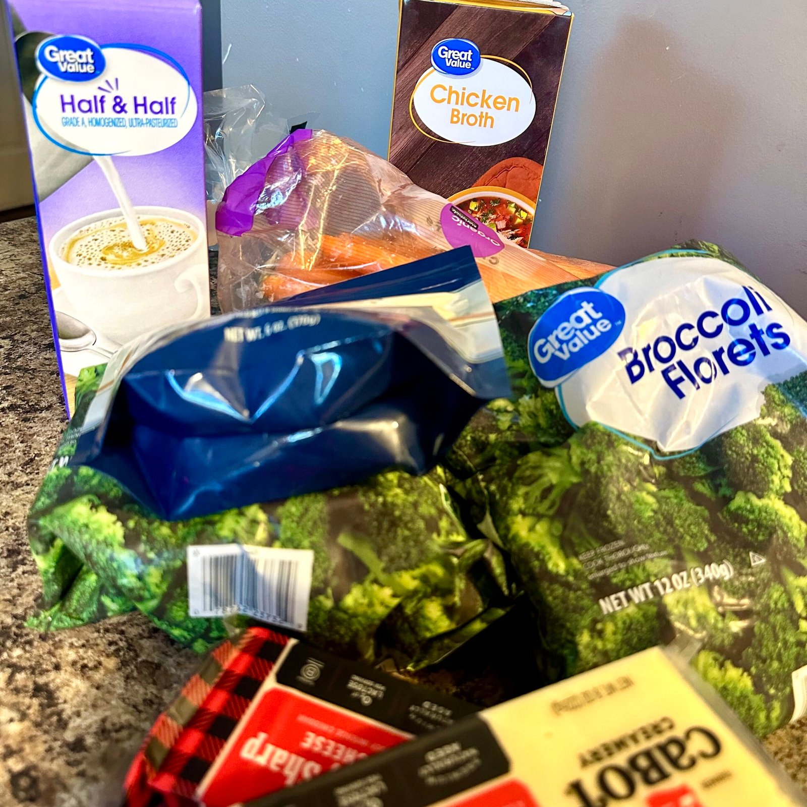 Ingredients for homemade broccoli cheddar soup, including broccoli florets, sharp cheddar cheese, carrots, half and half, and chicken broth on a kitchen counter.