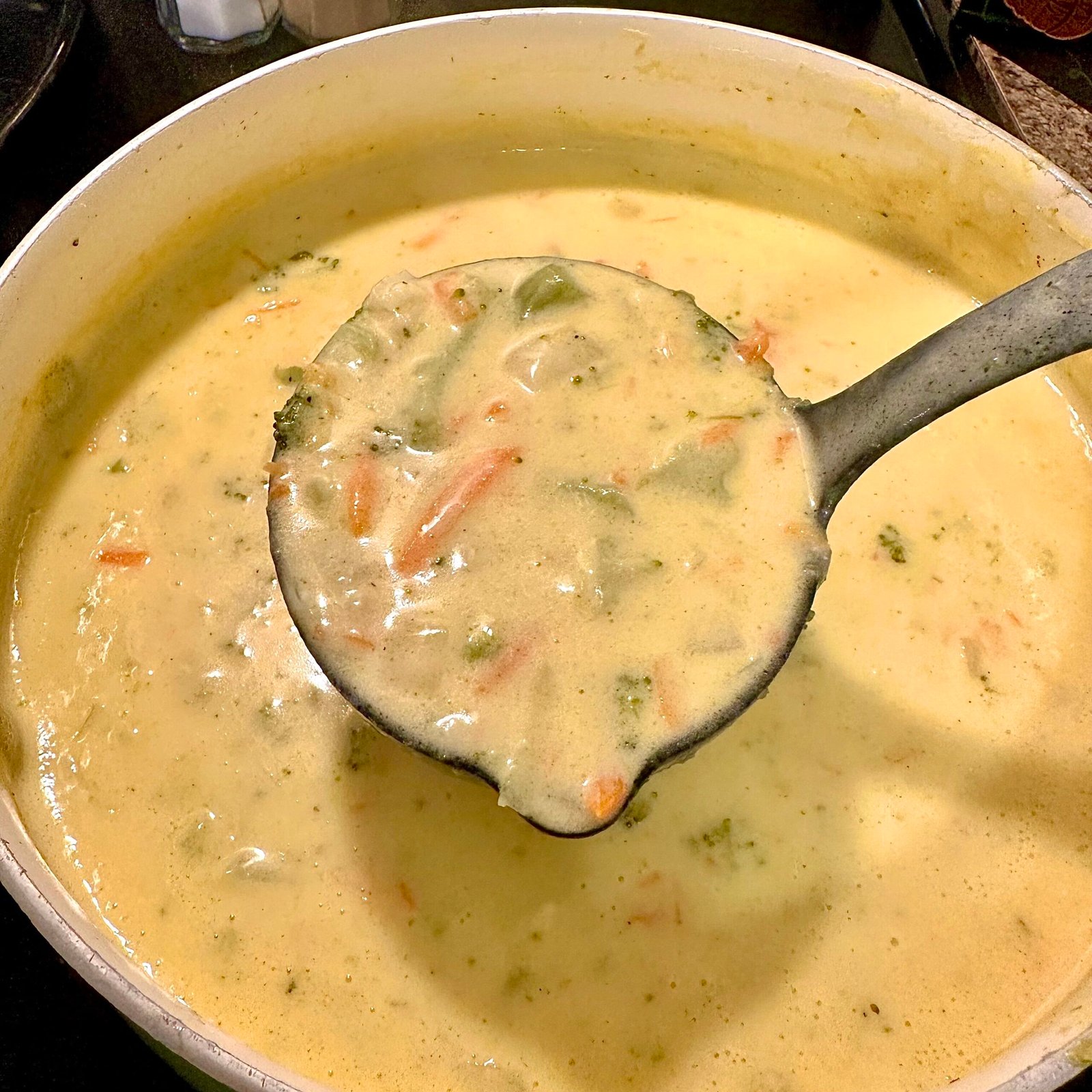 A ladle full of creamy homemade broccoli cheddar soup held over a pot, showing chunks of broccoli, carrots, and melted cheese.