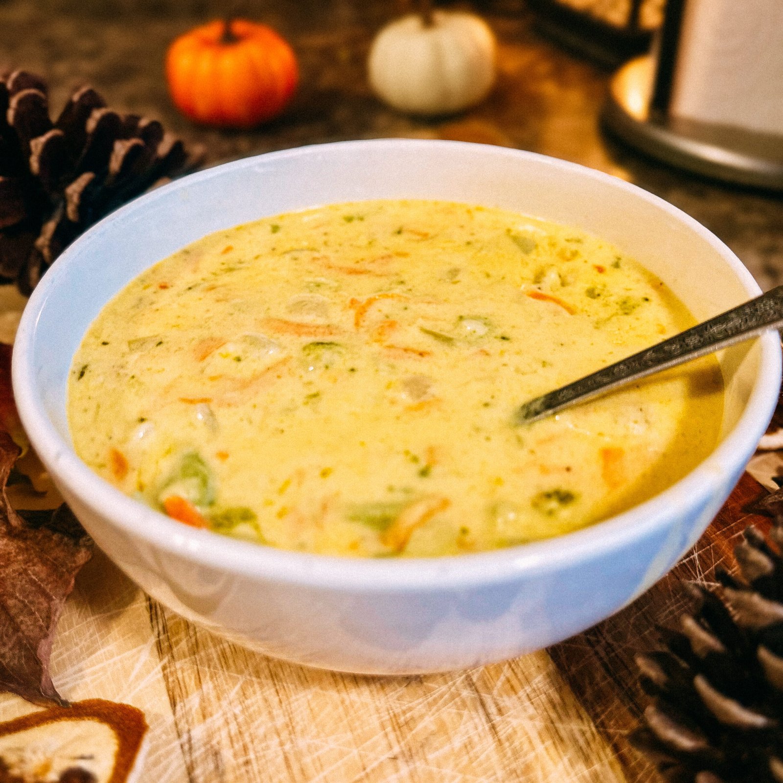 Bowl of creamy homemade broccoli cheddar soup surrounded by fall decor, pinecones, and mini pumpkins.